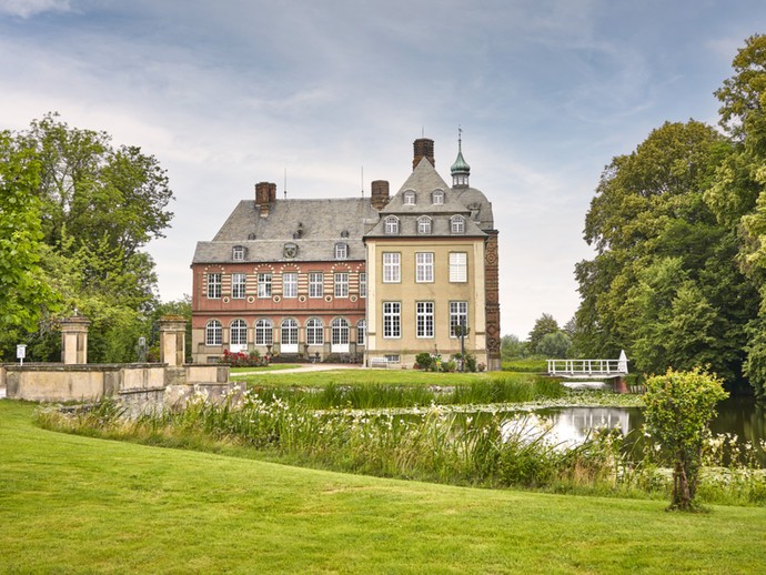 Blick über die Gräfte auf Schloss Hovestadt. Auf dem Wasser befinden sich Seerosen. Am Ufer befinden sich zahlreiche Pflanzen.